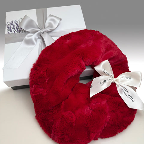Red fluffy neck pillow with a white bow and 'Sonoma' label next to a white gift box with a silver bow on a gray background.